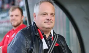 Prezes Polonii Warszawa w skórzanej kurtce i szaliku klubu, z uśmiechem na twarzy.