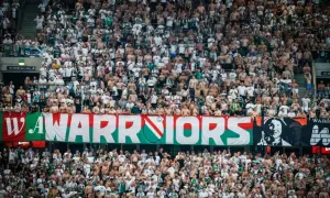 Tłum kibiców Legii Warszawa, z transparentem "WARRIORS" i herbem klubu. Wszyscy w biało-zielonych barwach, gotowi do dopingu.