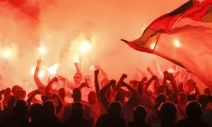 Tłum kibiców w czerwonym dymie z rac, z uniesionymi pięściami i flagą. Widać też sylwetki policjantów w hełmach.