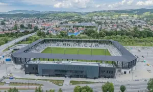 Nowoczesny stadion Sandecji z zieloną murawą i trybunami, otoczony miastem i wzgórzami.