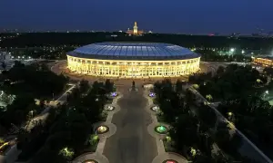 Oświetlony moskiewski stadion nocą, z aleją fontann i pomnikiem w tle.