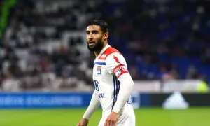 Piłkarz Nabil Fekir w białej koszulce z czerwonymi elementami, z opaską kapitańską na ramieniu, na tle trybun.