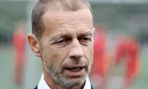 Aleksander Čeferin, prezes UEFA, w niebieskiej koszuli i granatowym swetrze, z rozmytym tłem piłkarzy.
