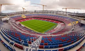 Widok na stadion w budowie, z czerwono-niebieskimi trybunami i zieloną murawą. W tle widoczne są dźwigi, sugerujące, że to jeden z największych stadionów w Polsce.