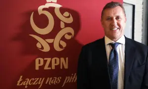 Uśmiechnięty prezes PZPN w garniturze stoi obok czerwonego banera z logo związku i hasłem "Łączy nas piłka".