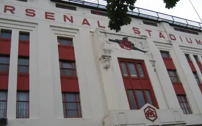 Stary stadion Arsenalu z czerwonymi oknami i napisem "ARSENAL STADIUM". Widoczny herb klubu i armata.