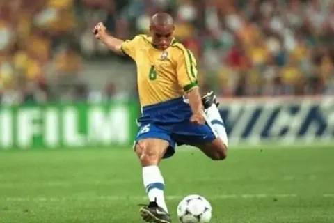 Roberto Carlos, z numerem 6 na koszulce, wykonuje potężne uderzenie piłki na boisku.