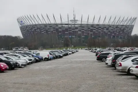 Duży parking przed Stadionem Narodowym w Warszawie. Widać rzędy samochodów i autobusów na błoniach stadionu.