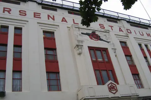 Stary stadion Arsenalu z czerwonymi oknami i napisem "ARSENAL STADIUM". Widoczny herb klubu i armata.