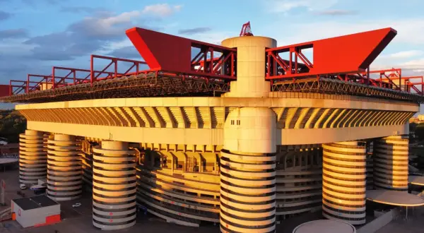 Stadion AC Milan w złotym świetle zachodzącego słońca, z charakterystycznymi czerwonymi elementami konstrukcji i betonowymi filarami.