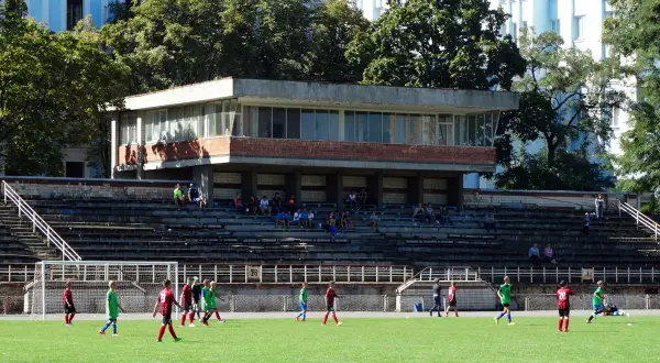 Młodzi piłkarze trenują na boisku. W tle trybuny i budynek, przypominający stadion Dynamo Kijów.