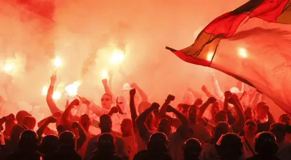 Tłum kibiców w czerwonym dymie z rac, z uniesionymi pięściami i flagą. Widać też sylwetki policjantów w hełmach.