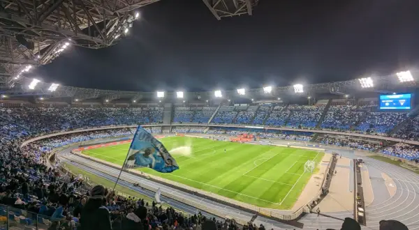 Widok na pełen kibiców stadion w Neapolu nocą. Trybuny wypełnione niebieskimi barwami, na murawie zielona trawa.