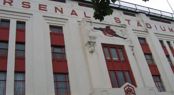 Stary stadion Arsenalu z czerwonymi oknami i napisem "ARSENAL STADIUM". Widoczny herb klubu i armata.