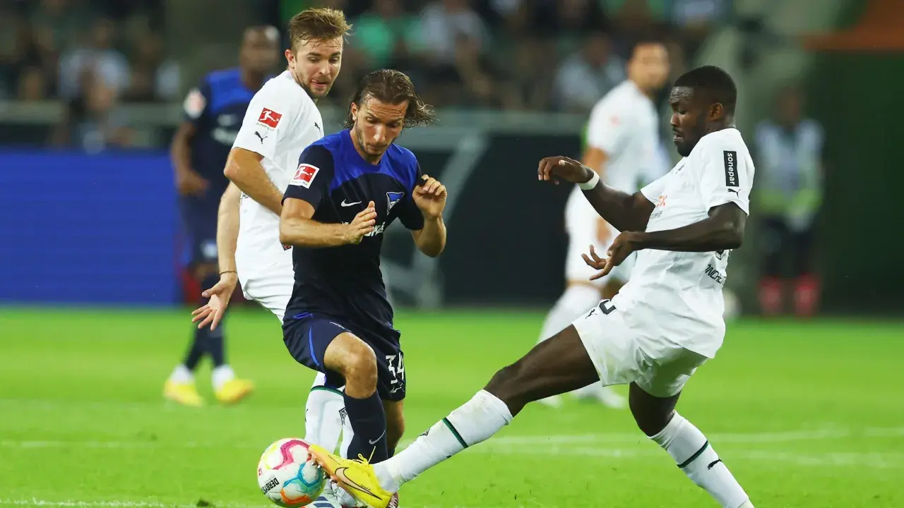 Piłkarze walczą o piłkę na boisku, jeden z nich w białej koszulce z logo Bundesligi, być może to przyszli bundesliga mistrzowie.