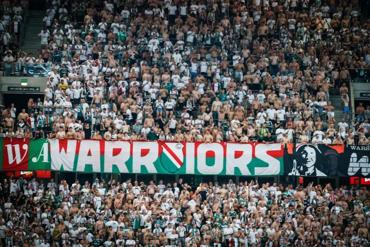 Tłum kibiców Legii Warszawa, z transparentem "WARRIORS" i herbem klubu. Wszyscy w biało-zielonych barwach, gotowi do dopingu.