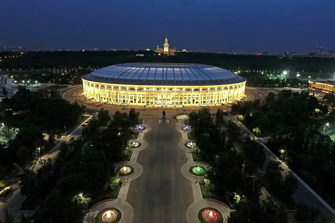 Oświetlony moskiewski stadion nocą, z aleją fontann i pomnikiem w tle.