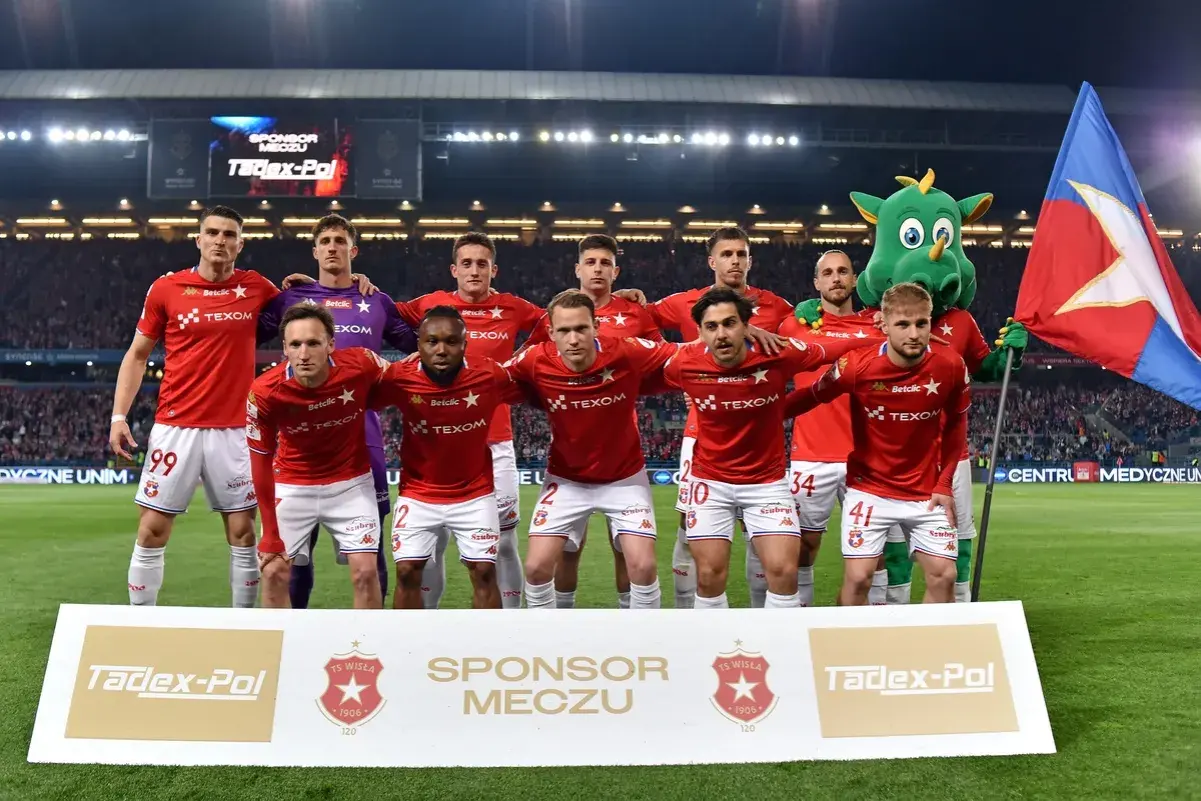 Piłkarze Wisły Kraków w czerwonych strojach, z maskotką i flagą. Najnowsze wisła kraków aktualności.