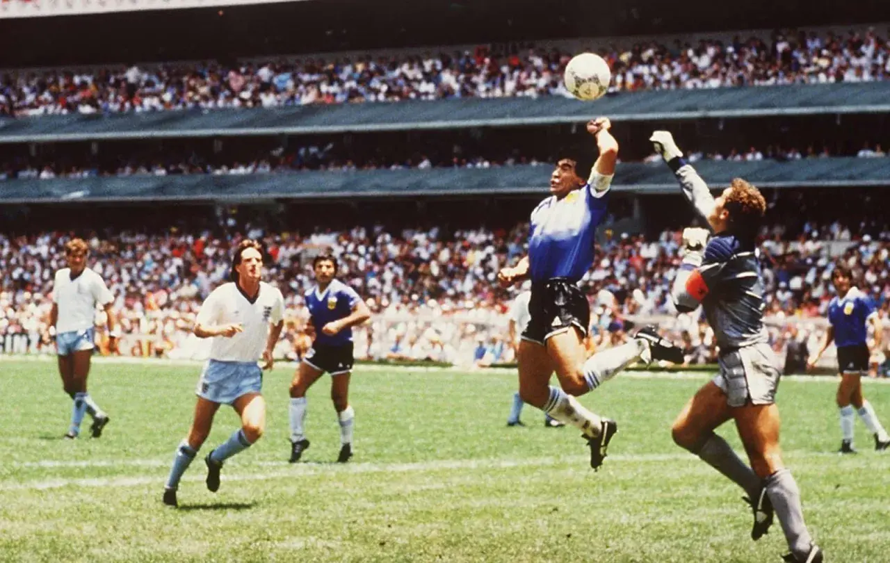 Diego Maradona, w słynnym meczu, używa "ręka boga", by zdobyć bramkę. Piłkarze w białych strojach patrzą z niedowierzaniem.