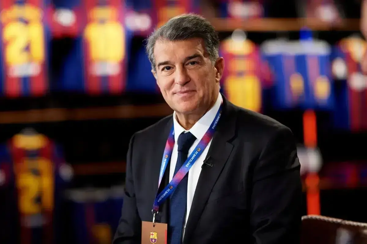 Prezes FC Barcelona, Joan Laporta, w garniturze, z identyfikatorem klubu na szyi, na tle klubowych koszulek.