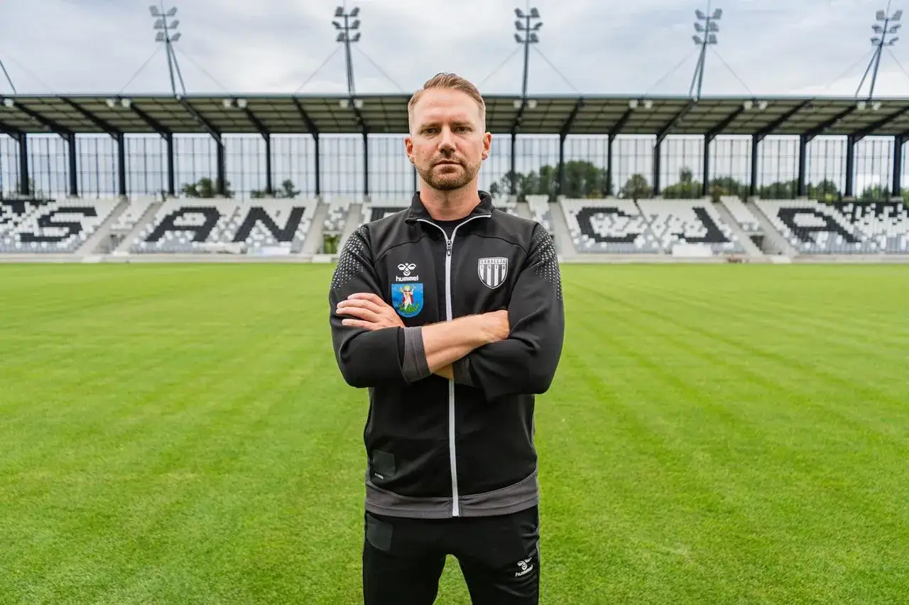 Trener z licencją UEFA Pro stoi na boisku piłkarskim przed trybunami.