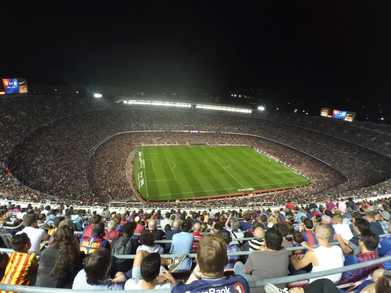 Nocny mecz na stadionie Barcelony. Tysiące kibiców na trybunach, zielona murawa oświetlona jupiterami.