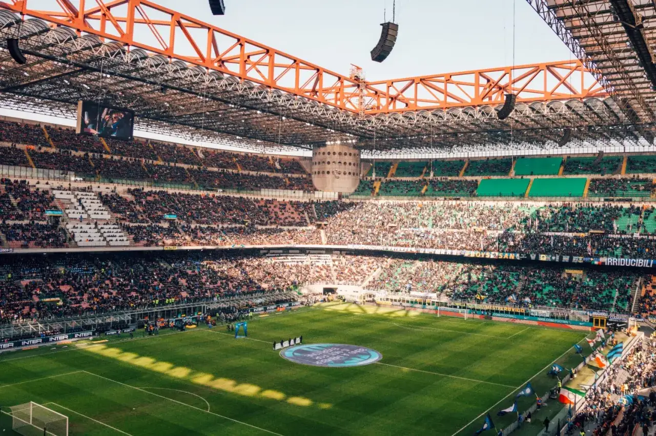 Stadion pełen kibiców podczas meczu włoskiej ligi. Zielona murawa, trybuny wypełnione ludźmi, a nad nimi stalowa konstrukcja dachu.