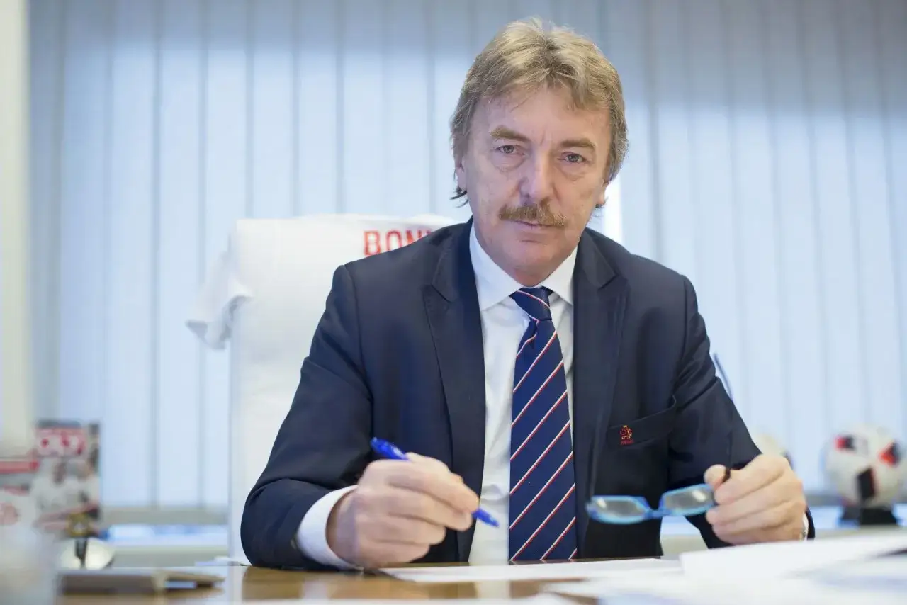Prezes PZPN, Zbigniew Boniek, w garniturze siedzi przy biurku, trzymając długopis i okulary.