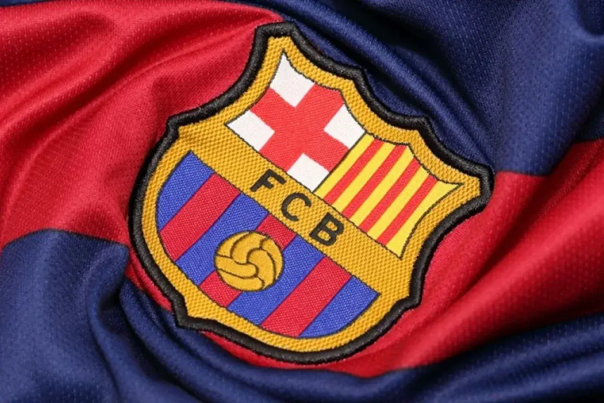 Herb FC Barcelona, znany jako logo Barcy, na tle klubowej koszulki w bordowo-granatowe pasy.