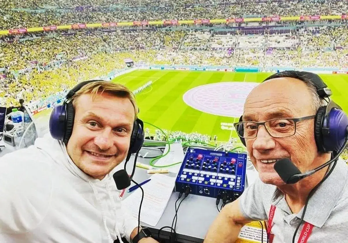 Dwóch komentatorów sportowych w słuchawkach z mikrofonami przed stadionem pełnym kibiców.