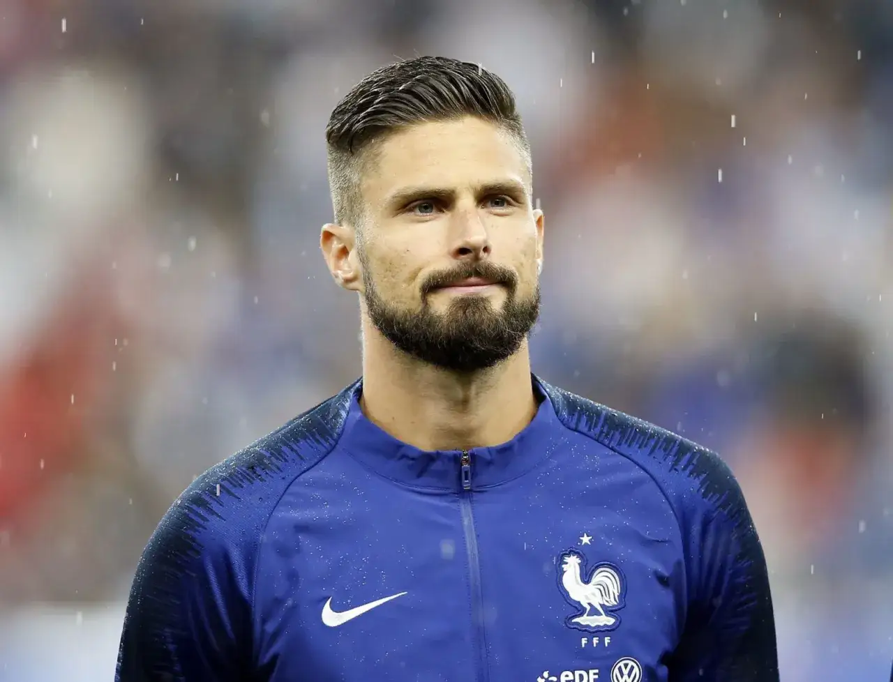 Olivier Giroud w stylowej fryzurze piłkarskiej, w deszczu, w niebieskiej bluzie reprezentacji Francji.