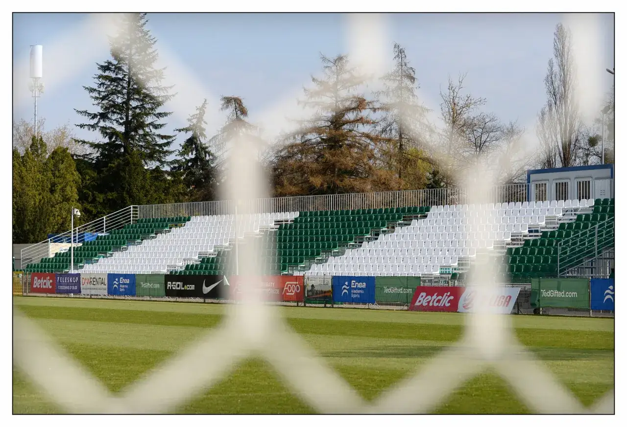 Puste trybuny stadionu Warty Poznań, zielono-białe krzesełka i boisko. W tle drzewa i maszt telekomunikacyjny.