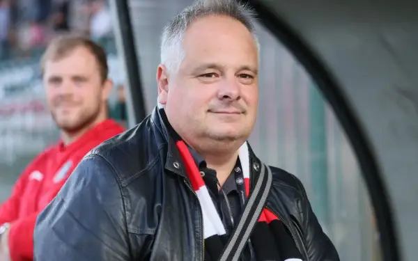 Prezes Polonii Warszawa w skórzanej kurtce i szaliku klubu, z uśmiechem na twarzy.