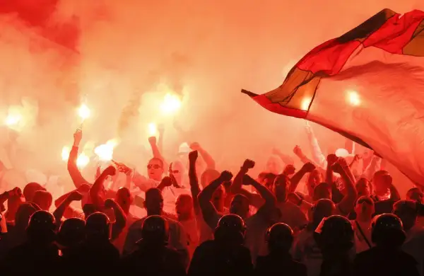 Tłum kibiców w czerwonym dymie z rac, z uniesionymi pięściami i flagą. Widać też sylwetki policjantów w hełmach.