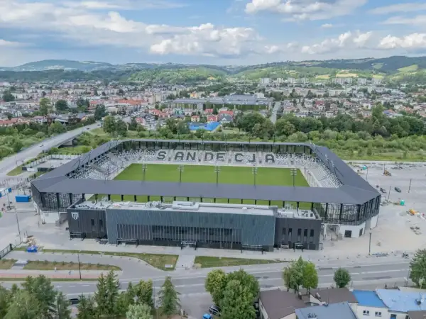 Nowoczesny stadion Sandecji z zieloną murawą i trybunami, otoczony miastem i wzgórzami.