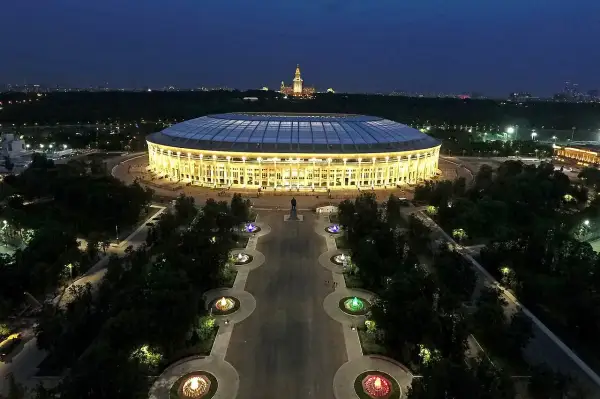 Oświetlony moskiewski stadion nocą, z aleją fontann i pomnikiem w tle.