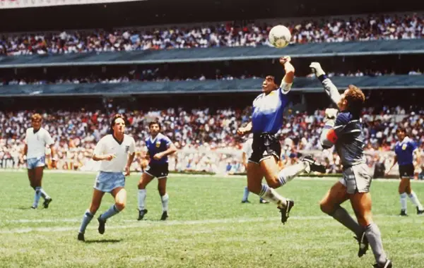 Diego Maradona, w słynnym meczu, używa "ręka boga", by zdobyć bramkę. Piłkarze w białych strojach patrzą z niedowierzaniem.