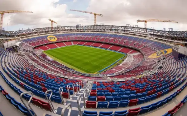 Widok na stadion w budowie, z czerwono-niebieskimi trybunami i zieloną murawą. W tle widoczne są dźwigi, sugerujące, że to jeden z największych stadionów w Polsce.